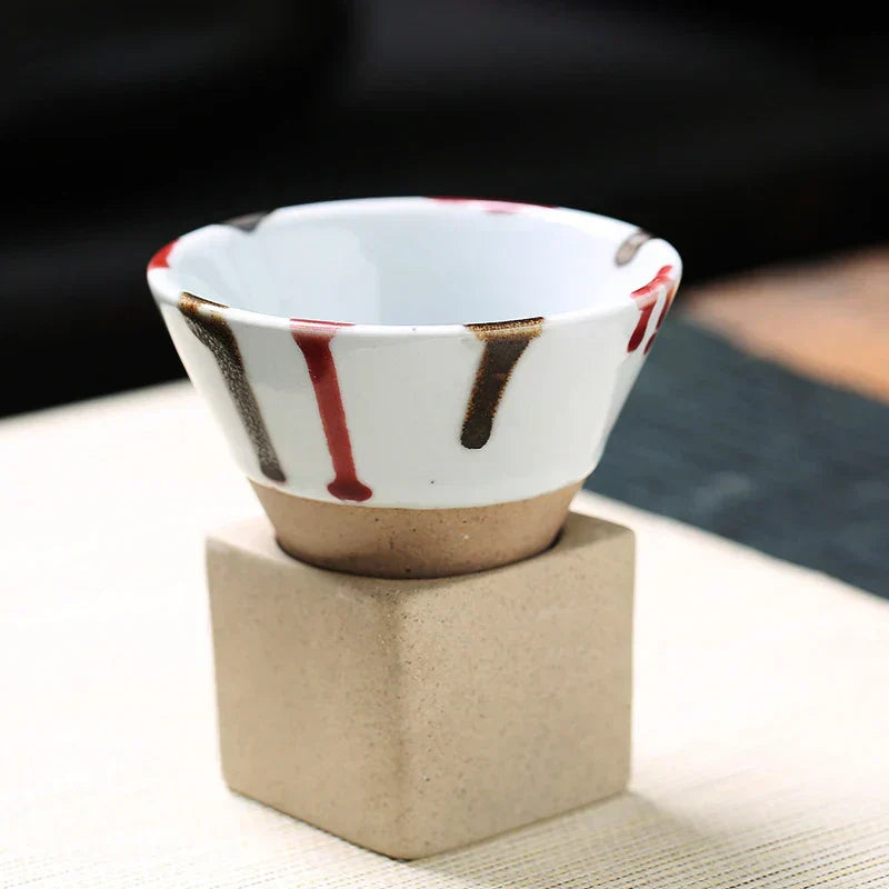 Weißer Keramikbecher mit braunen und roten Streifen, auf beigem Würfel, modernes Design.