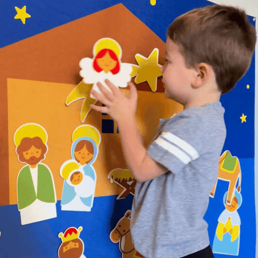 Kleinkind spielt mit Krippenfiguren aus Filz, Weihnachtsdekoration, blau-gelber Hintergrund.