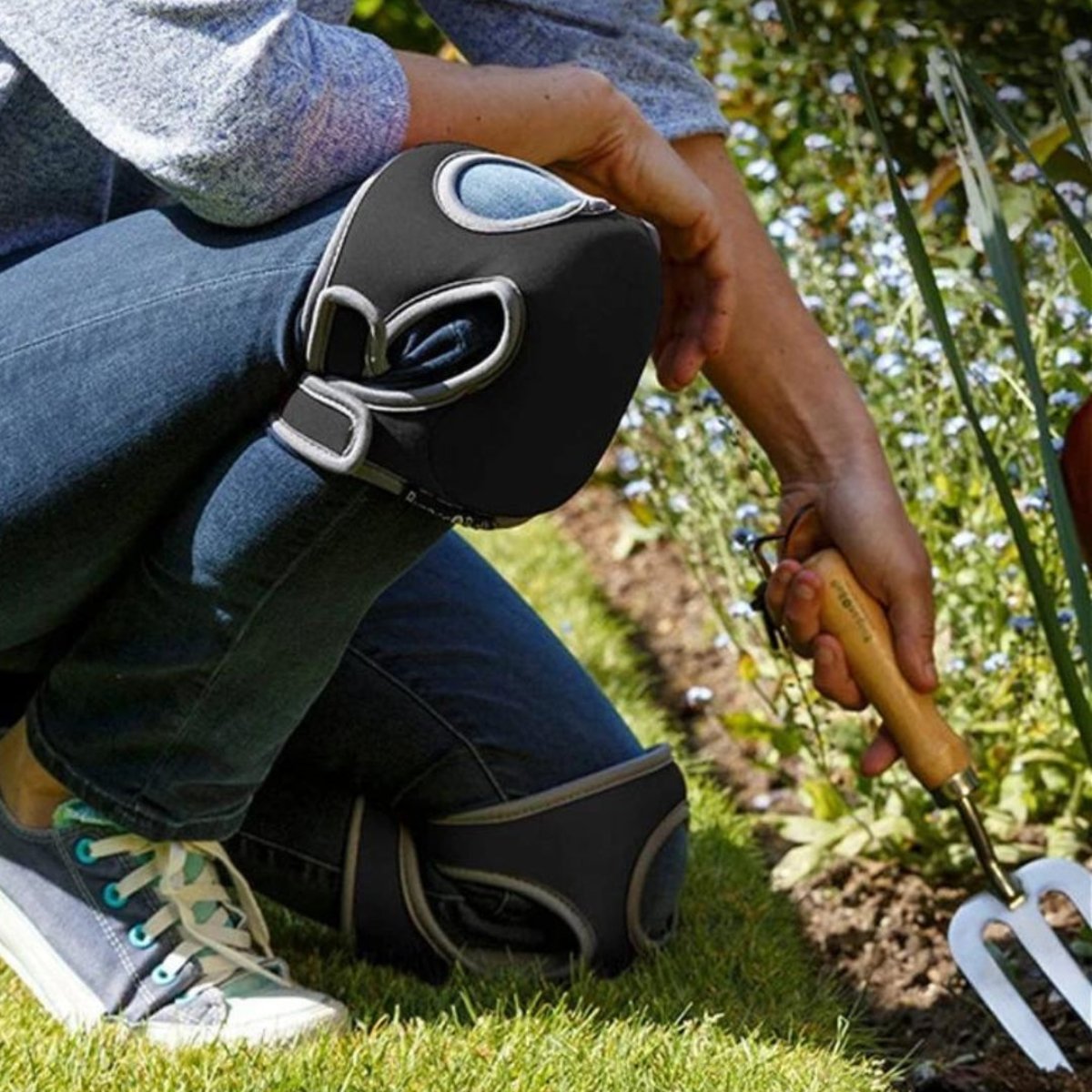 Person mit schwarzen Knieschonern beim Gärtnern, hält Gartenwerkzeug, Blumenbeet im Hintergrund.