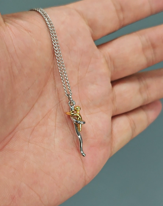 Der Anhänger mit den beiden umarmenden Figuren in Silber- und Goldton liegt samt Kette auf einer offenen Handfläche und zeigt die Größe. 7