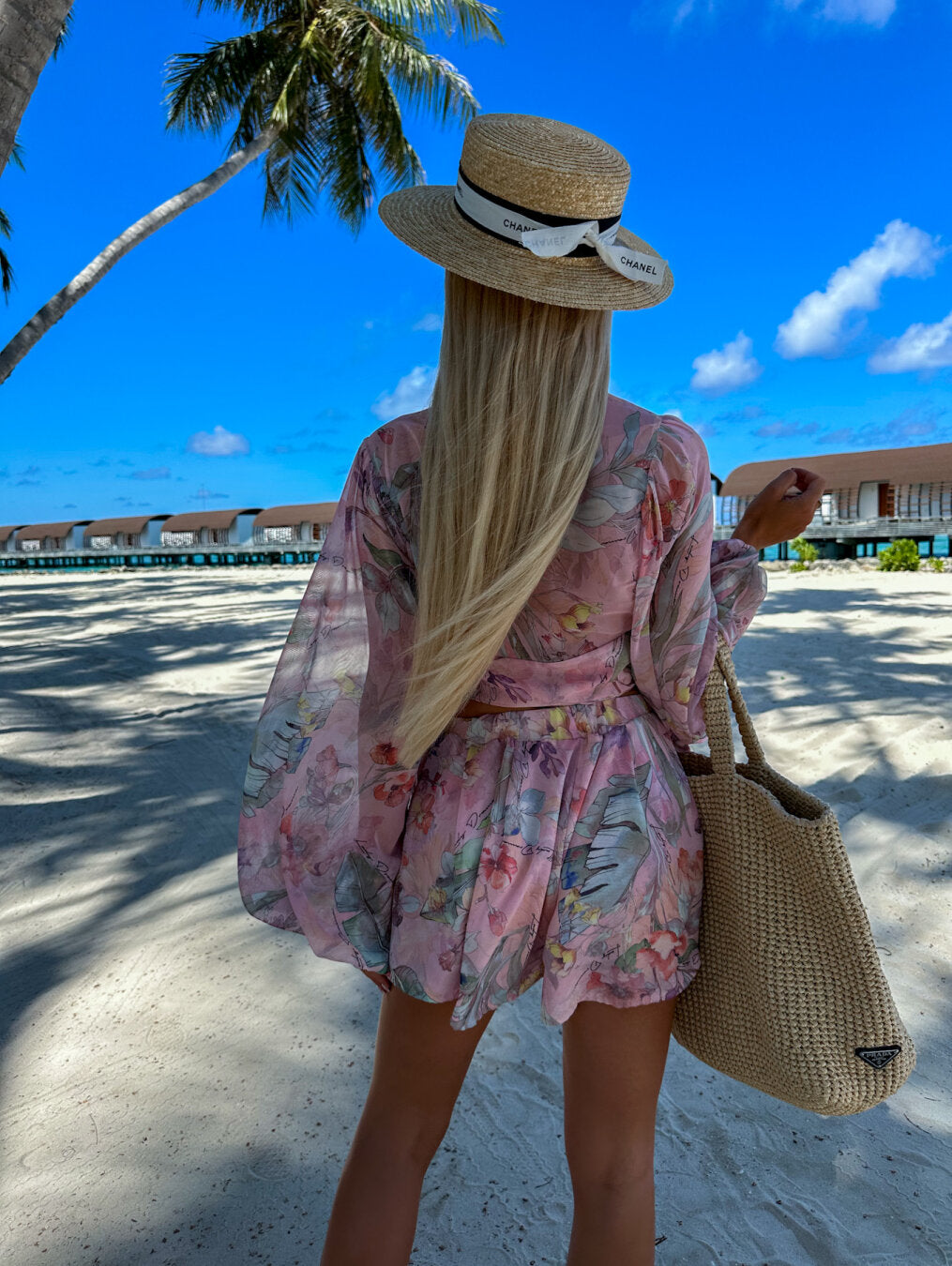 Frau im floralen Sommerkleid und Strohhut am Strand, mit Palmen und blauen Himmel im Hintergrund.