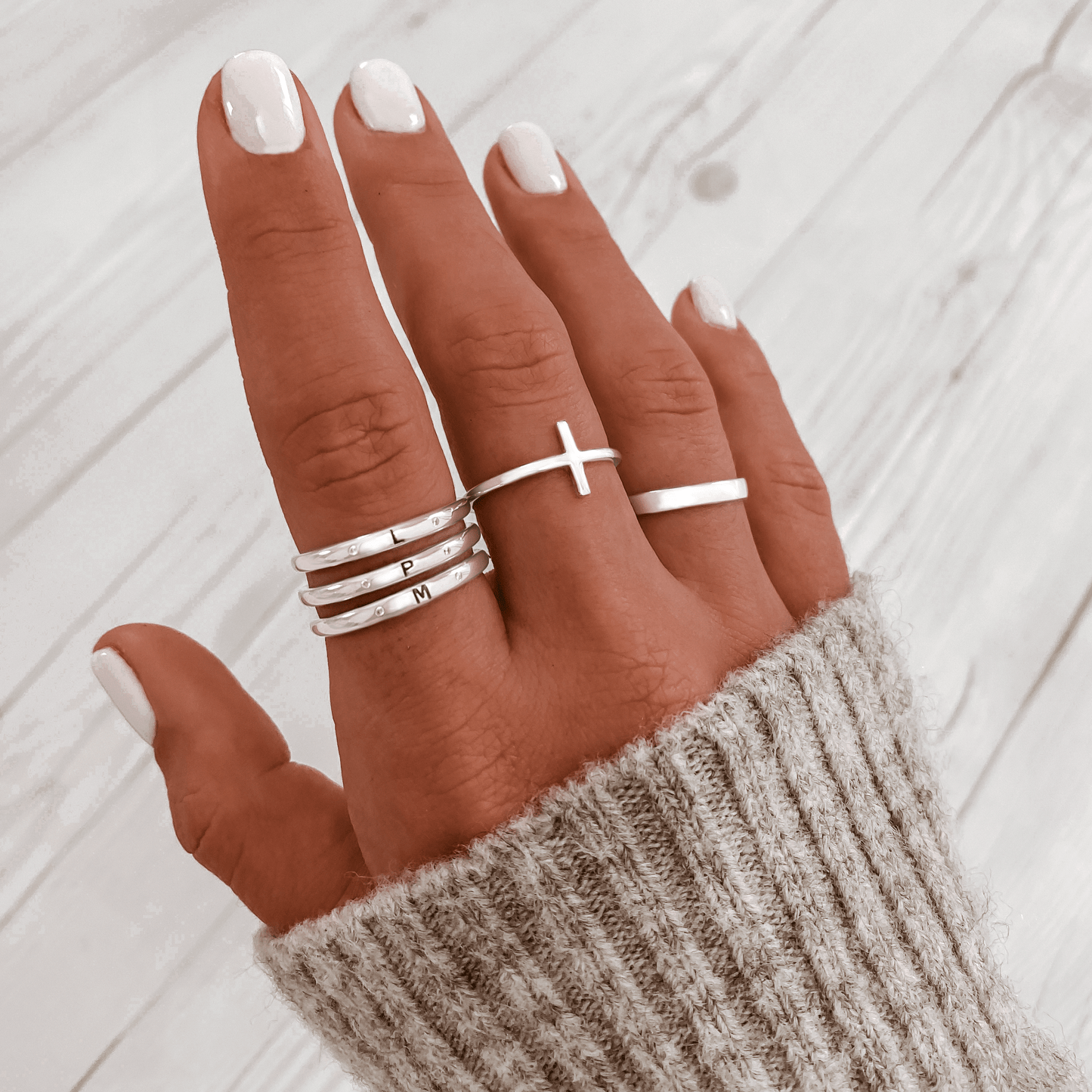 Hand mit silbernen Ringen, Kreuzring, Buchstabenringe, grauer Strickpullover, Nagellack.