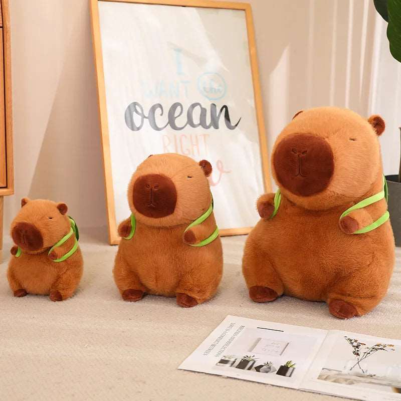Drei braune Capybara-Plüschtiere mit grünen Schals in verschiedenen Größen auf Teppich.