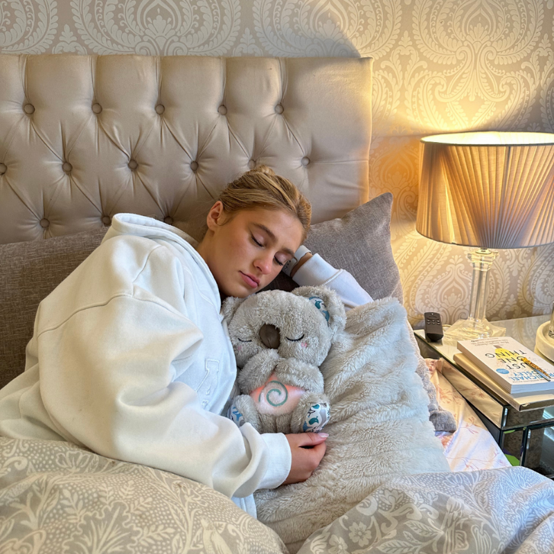 Frau schläft im Bett mit Plüsch-Koala, beige Bettwäsche, Nachttischlampe, gemütliches Schlafzimmer.