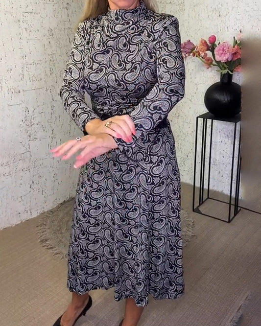 Frau in langem, gemustertem Kleid mit Paisley-Muster, Blumen im Hintergrund, elegante Mode.