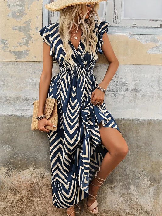 Frau in blau-weißem Maxikleid mit Zickzackmuster, Strohhut und Clutch vor rustikaler Wand.