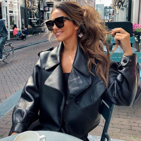 Frau in schwarzer Lederjacke und Sonnenbrille sitzt draußen, lächelt, hält Haare hoch.