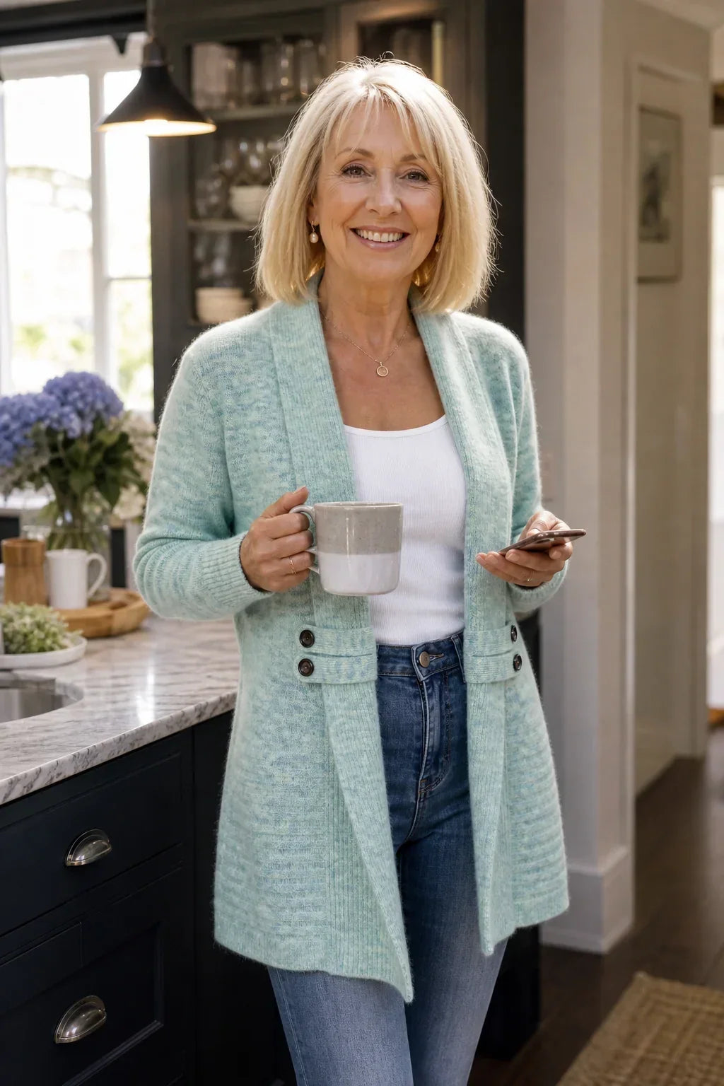 Ältere Frau in mintgrünem Cardigan mit Schalkragen, lächelnd mit Tasse und Smartphone in der Küche 4