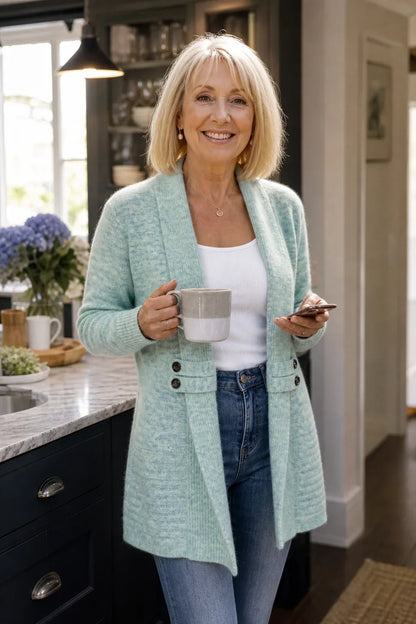 Ältere Frau in mintgrünem Cardigan mit Schalkragen, lächelnd mit Tasse und Smartphone in der Küche 4