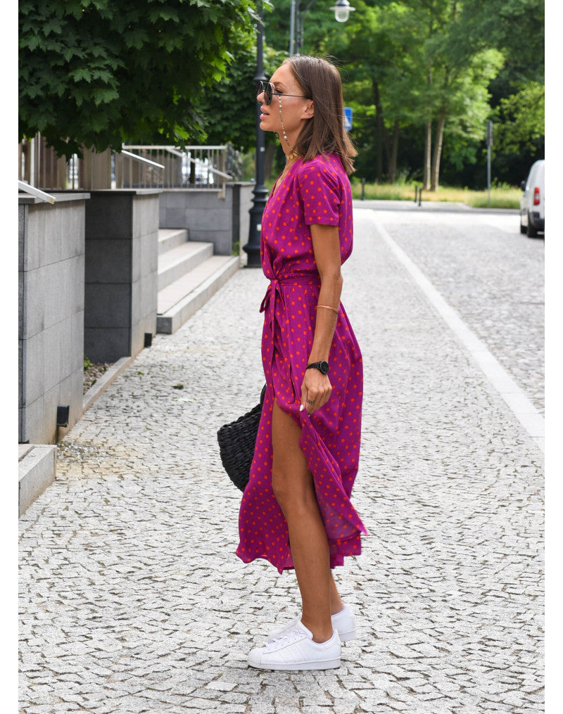 Frau in pinkem Sommerkleid mit Punkten, trägt weiße Sneaker, schwarze Tasche, Sonnenbrille.