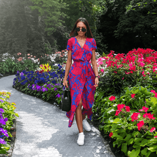 Frau in rotem Sommerkleid mit blauem Muster spaziert im blühenden Garten, trägt Sonnenbrille.