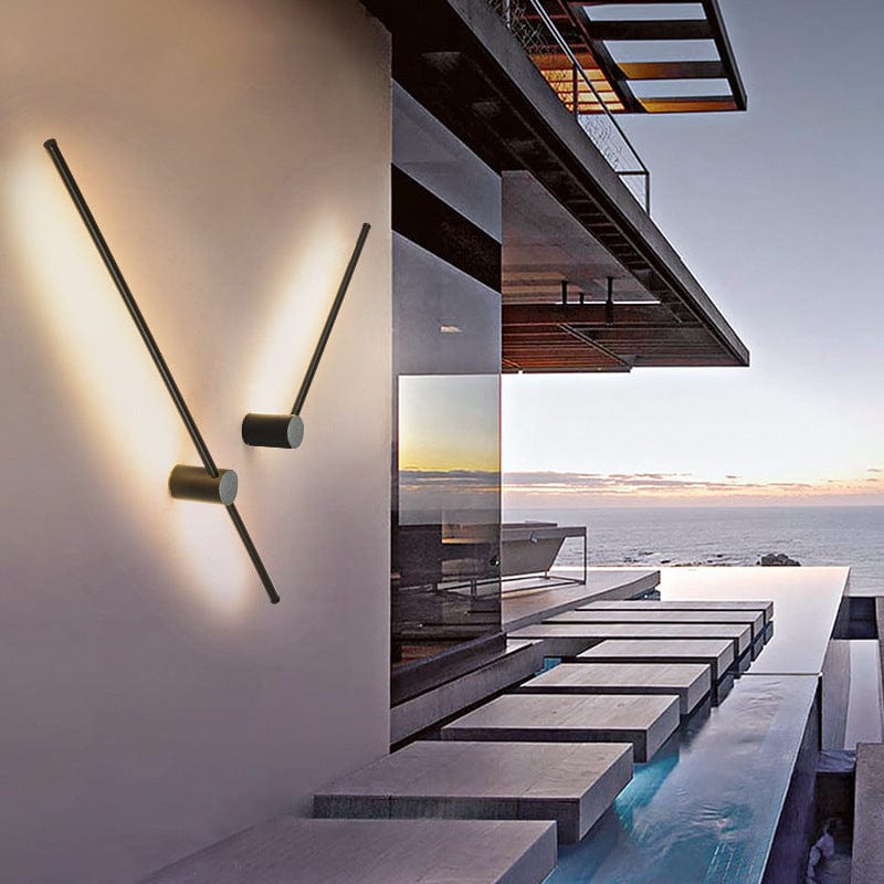 Zwei schräg gedrehte schwarze Stab-Wandlampen strahlen warmes Licht an eine Hauswand, im Hintergrund Meer und Pool bei Sonnenuntergang. 2