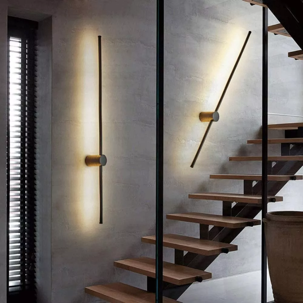Eine senkrechte und eine schräg gestellte Stab-Wandlampe beleuchten eine Betonwand neben einer Treppe mit Holzstufen im Innenbereich. 4