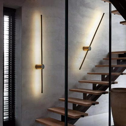 Eine senkrechte und eine schräg gestellte Stab-Wandlampe beleuchten eine Betonwand neben einer Treppe mit Holzstufen im Innenbereich. 4