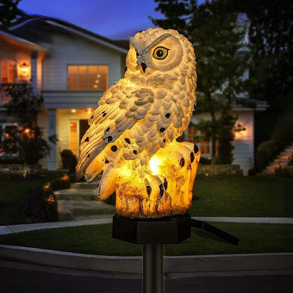 Weiße Eule mit dunklen Punkten leuchtet warm auf einem Baumstumpf-Sockel, im Hintergrund ein beleuchtetes Wohnhaus am Abend. 2