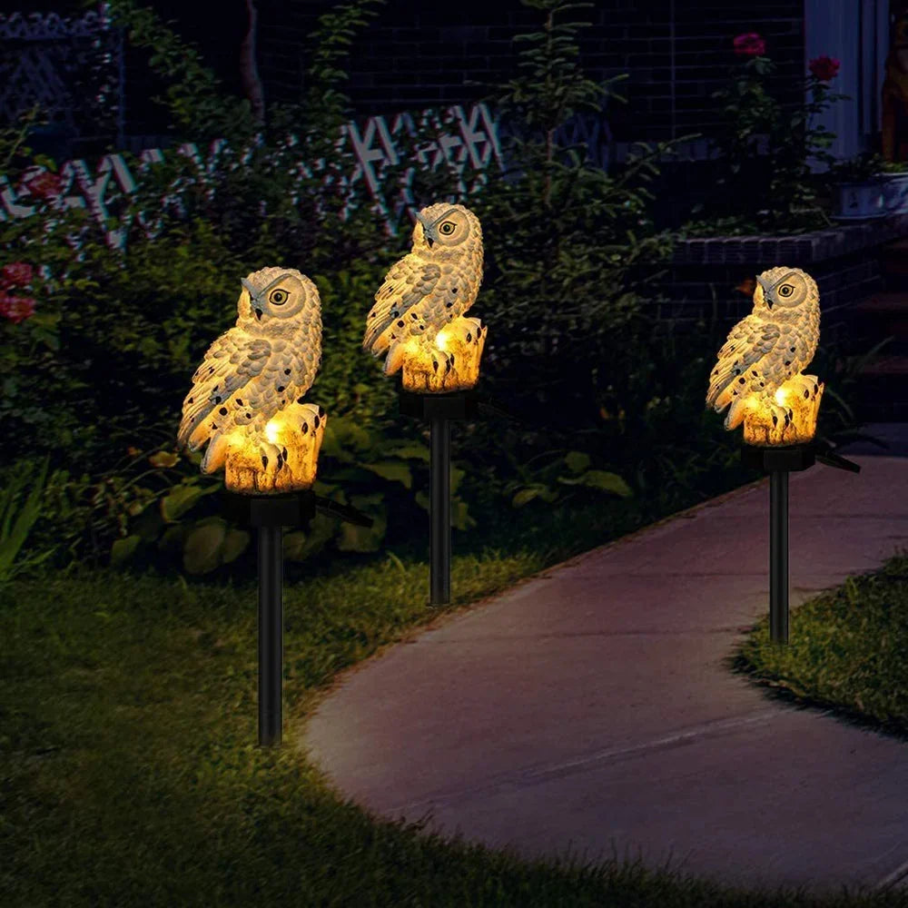 Drei beleuchtete Eulen-Gartenstecker säumen nachts einen Gartenweg zwischen Büschen und Rosen. 3