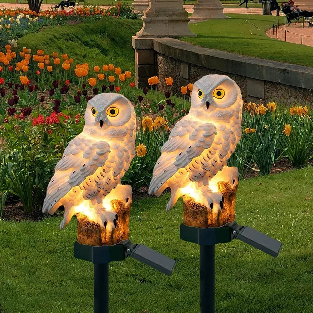 Zwei leuchtende weiße Eulen auf Erdspießen mit Solarpanel im Park vor blühenden Tulpen und Narzissen in der Abenddämmerung. 1