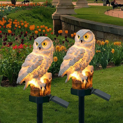 Zwei leuchtende weiße Eulen auf Erdspießen mit Solarpanel im Park vor blühenden Tulpen und Narzissen in der Abenddämmerung. 1