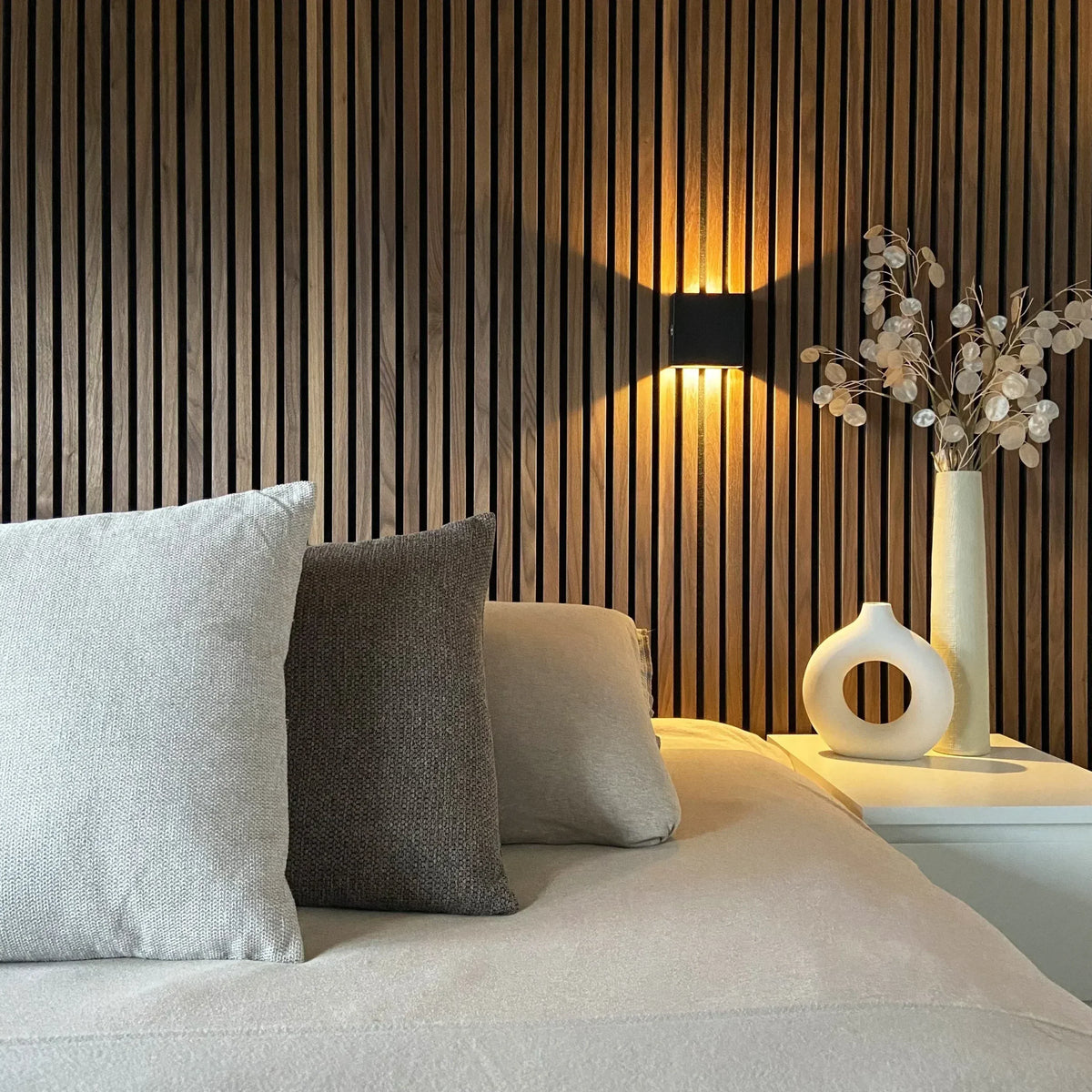 Schwarze Würfelleuchte vor einer Wand aus Holzlamellen über einem Bett mit Kissen und weißen Vasen auf dem Nachttisch. 5