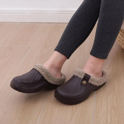 Person in dunkelgrauer Leggings trägt braune Clogs mit Plüschfutter auf hellem Holzboden, sitzend fotografiert. 7
