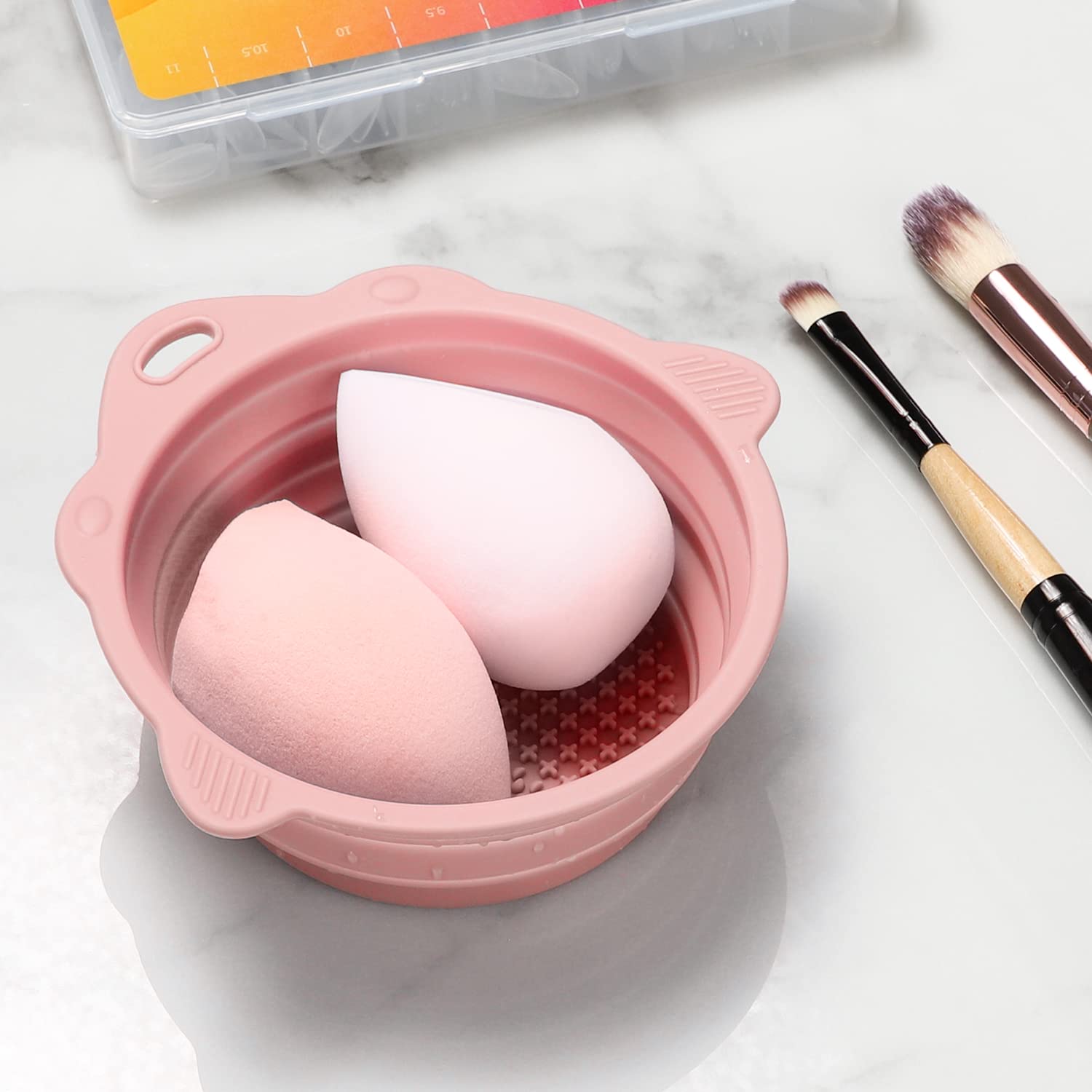 Rosa Silikon-Reinigungsbehälter mit Make-up-Schwämmen und Pinseln auf Marmortisch.