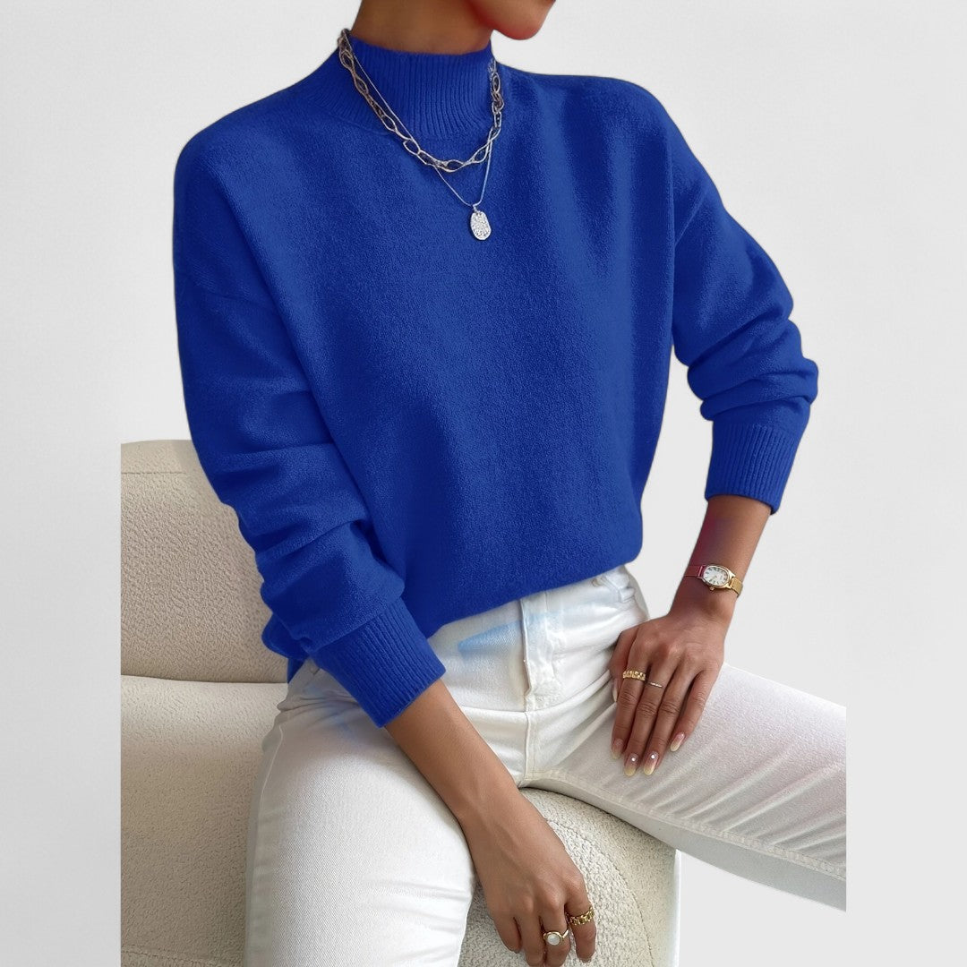 Pullover in kräftigem Blau mit Stehkragen am Model, dazu weiße Jeans, silberne Halskette und goldene Uhr, sitzend vor hellem Hintergrund. 11
