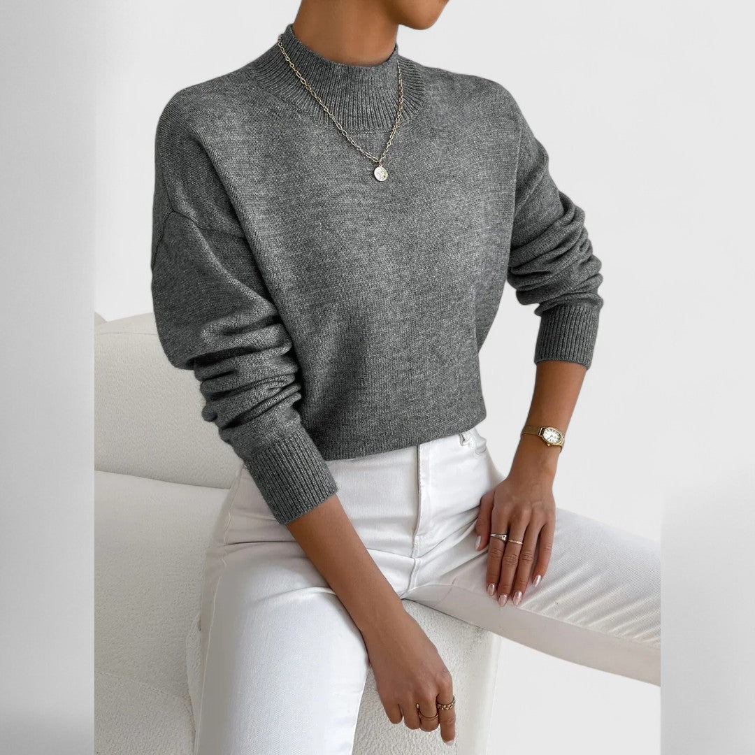 Grauer Pullover mit Stehkragen und Rippbündchen am sitzenden Model, kombiniert mit weißer Jeans, feiner Kette und goldener Armbanduhr. 12