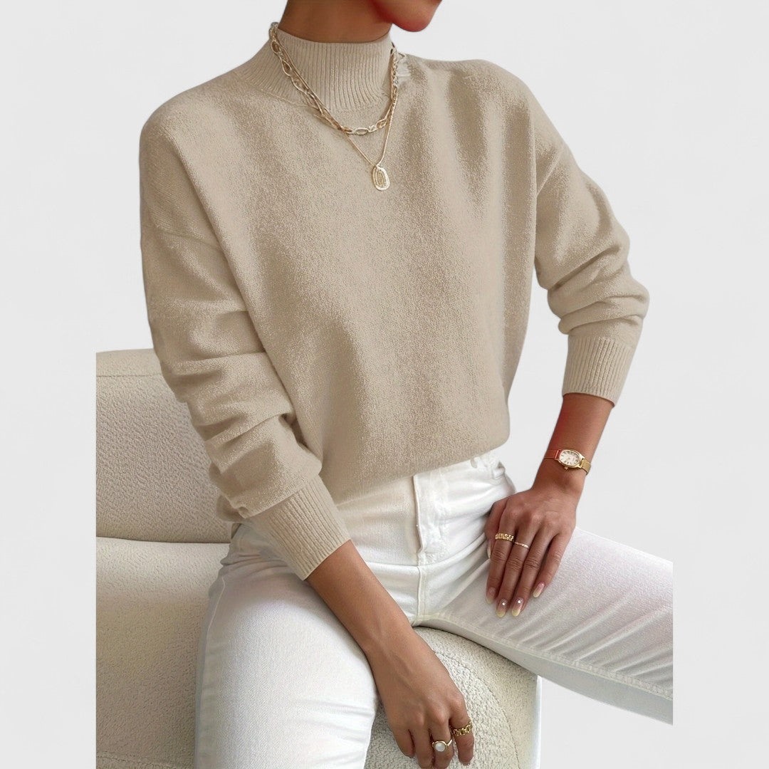 Beigefarbener Pullover mit gerippten Bündchen am Model, kombiniert mit weißer Jeans und goldenen Halsketten auf cremefarbenem Sessel. 7