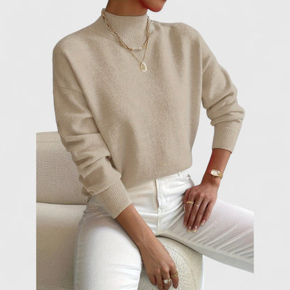 Beigefarbener Pullover mit gerippten Bündchen am Model, kombiniert mit weißer Jeans und goldenen Halsketten auf cremefarbenem Sessel. 7