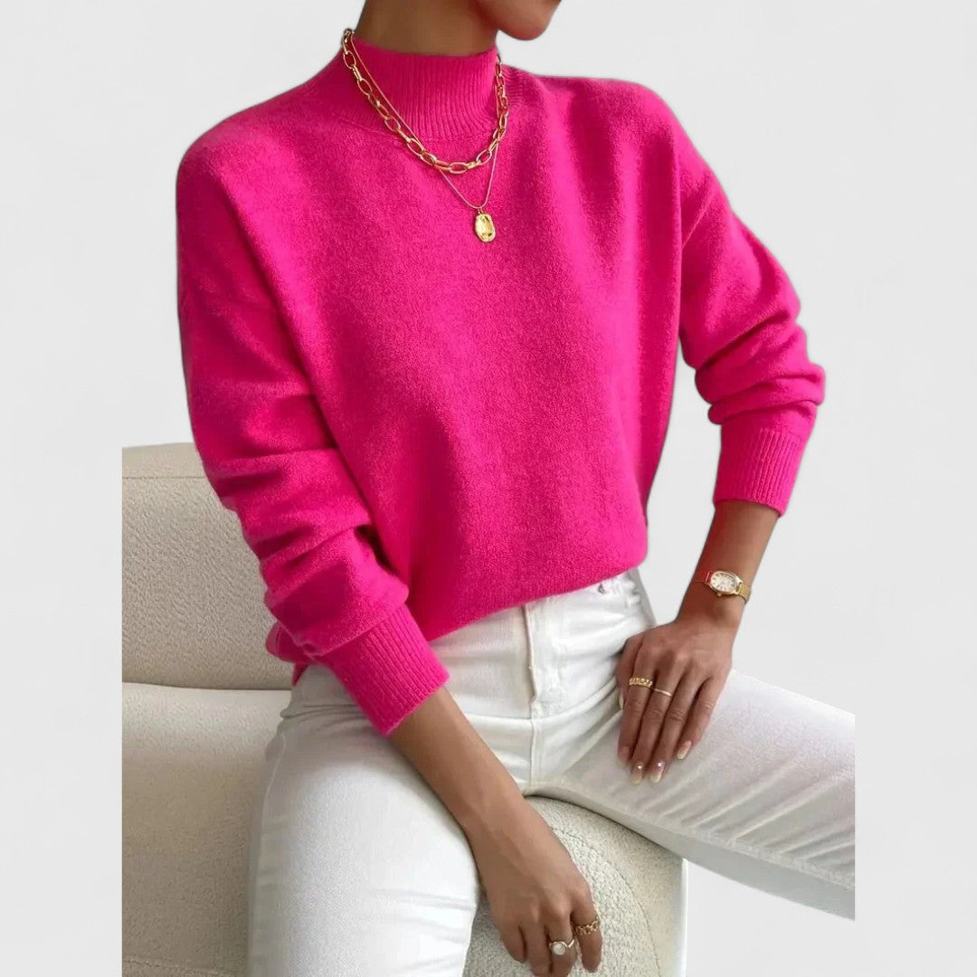 Pullover in leuchtendem Pink mit Stehkragen am sitzenden Model, dazu weiße Jeans, goldene Ketten, Ringe und eine Armbanduhr. 9