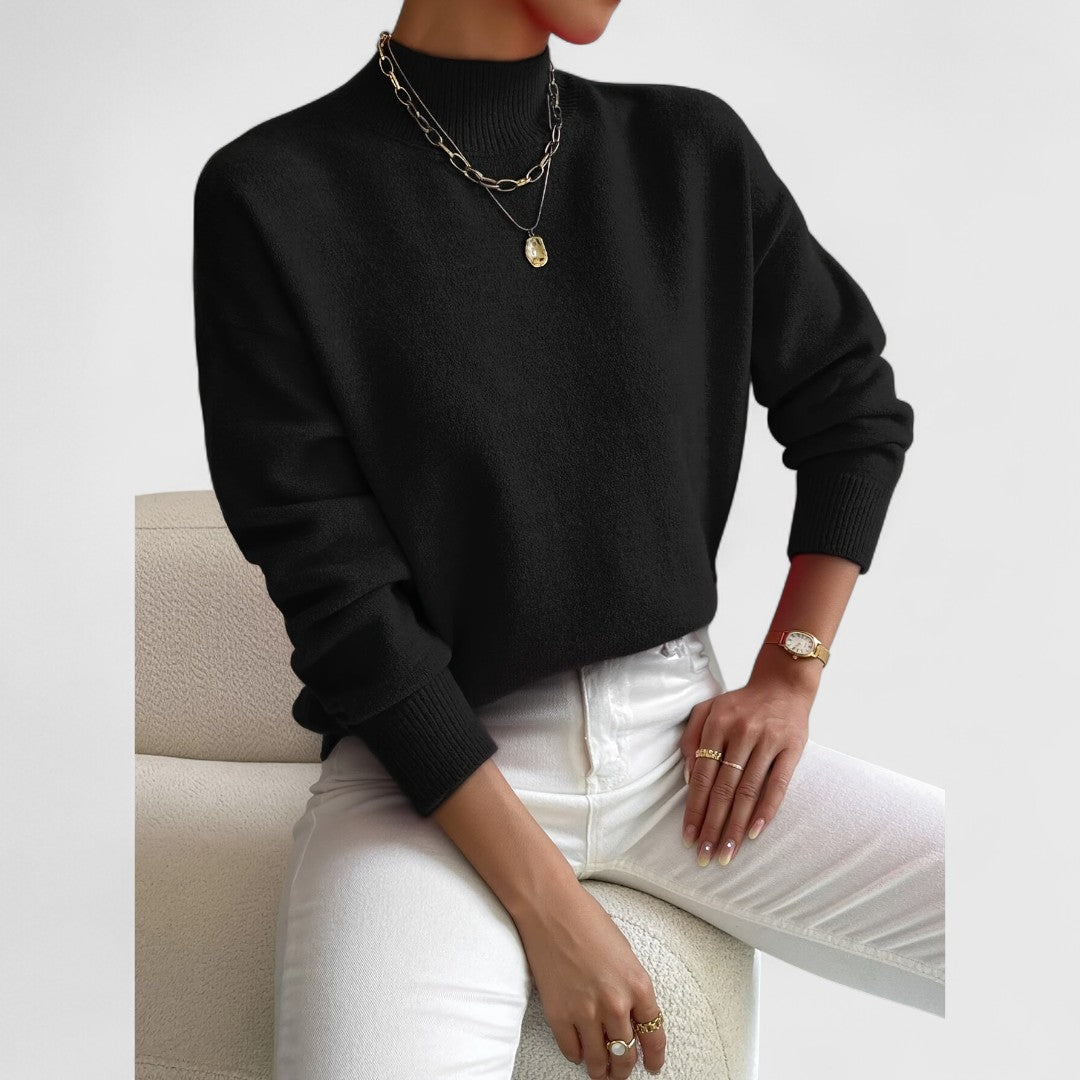 Schwarzer Pullover mit Stehkragen und Rippbündchen am Model, kombiniert mit weißer Jeans und goldenem Schmuck, sitzend auf einem hellen Sessel. 8