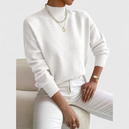 Weißer Pullover mit Stehkragen am Model, kombiniert mit weißer Jeans und goldenem Schmuck, locker sitzend auf einem Bouclé-Sessel. 4