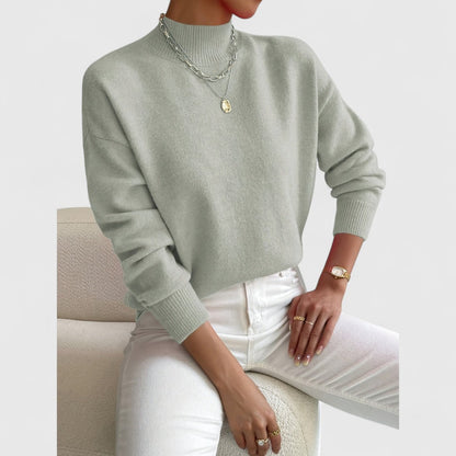 Graugrüner Pullover mit gerippten Bündchen am sitzenden Model, kombiniert mit weißer Jeans, Goldring, Uhr und mehrlagiger Halskette. 3