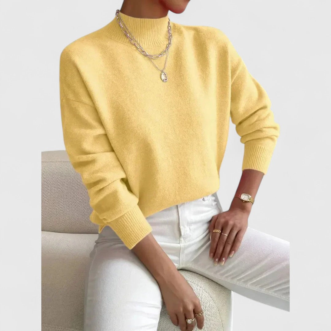 Pullover in sanftem Gelb mit Stehkragen am sitzenden Model, dazu weiße Jeans, Armbanduhr, Ringe und eine silberne Kette. 6