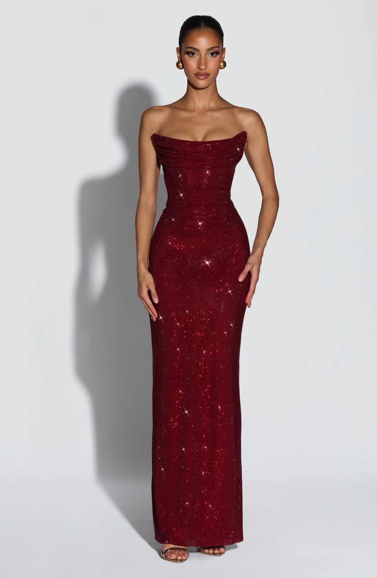 Model trägt das trägerlose Maxikleid in Dunkelrot mit Strassbesatz in der Frontansicht, mit drapiertem Mieder und goldenen Ohrringen. 1