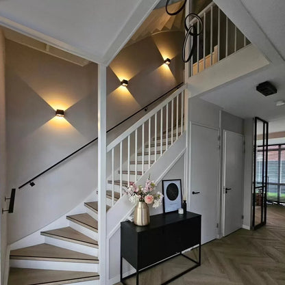 Drei schwarze Würfelleuchten entlang einer Treppe im Flur, darunter eine schwarze Kommode mit Blumenstrauß und Bilderrahmen. 7