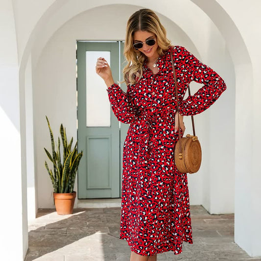 Frau in rotem Leopardenkleid mit Strohtasche, Sonnenbrille, steht vor weißem Torbogen.