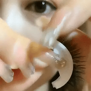 Nahaufnahme einer Hand, die Wimpern auf dem weißen Halter hält und sie an das Auge führt. 14
