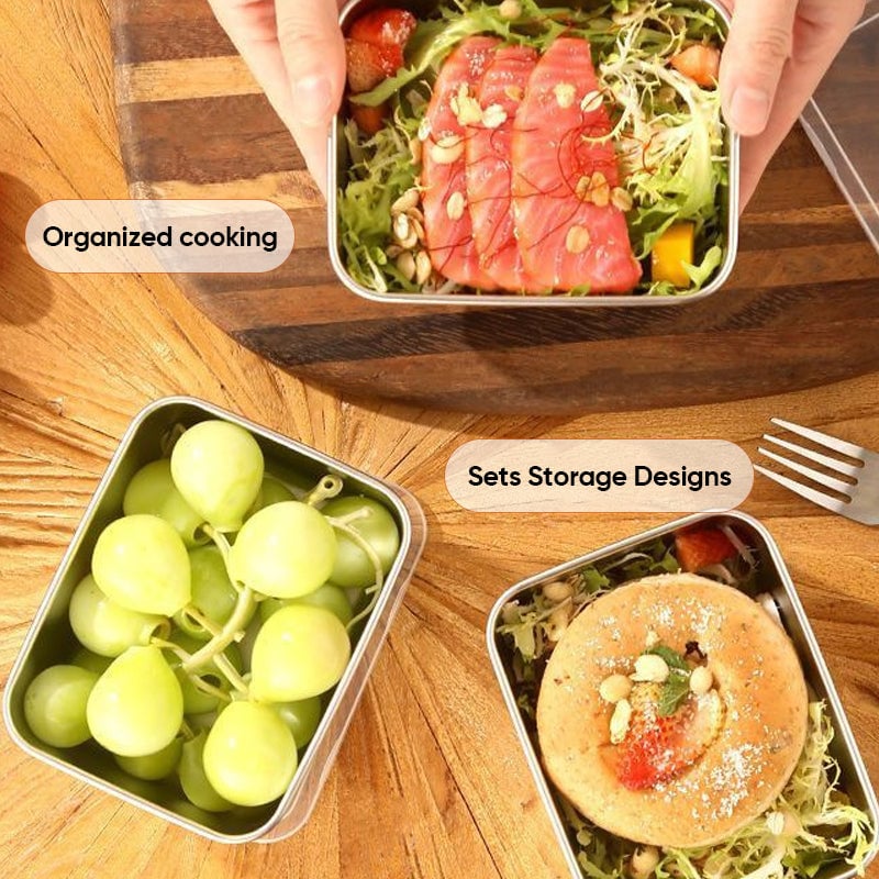 Draufsicht auf drei Edelstahlboxen auf Holztisch, gefüllt mit Lachs auf Salat, Trauben und einem Gebäck, daneben eine Gabel. 8