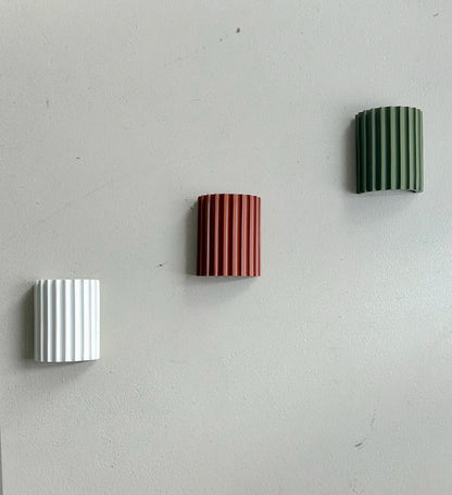 Drei gerillte Wandleuchten in Weiß, Terrakotta und Grün diagonal an einer grauen Wand montiert, ausgeschaltet 12