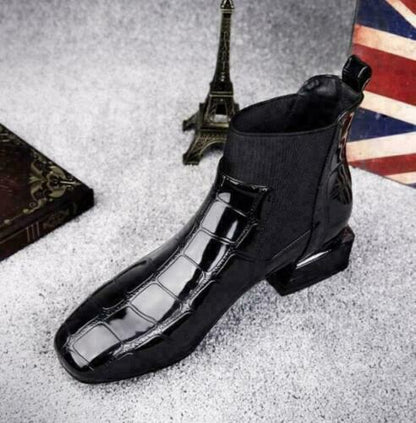 Schwarzer Chelsea-Stiefel mit eckiger Spitze und Krokooptik von schräg oben, daneben Eiffelturm-Figur und ein dunkles Buch. 5