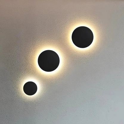 Drei schwarze runde Wandleuchten in unterschiedlichen Größen, diagonal angeordnet, mit warmem Lichtschein rund um die Scheibe. 2
