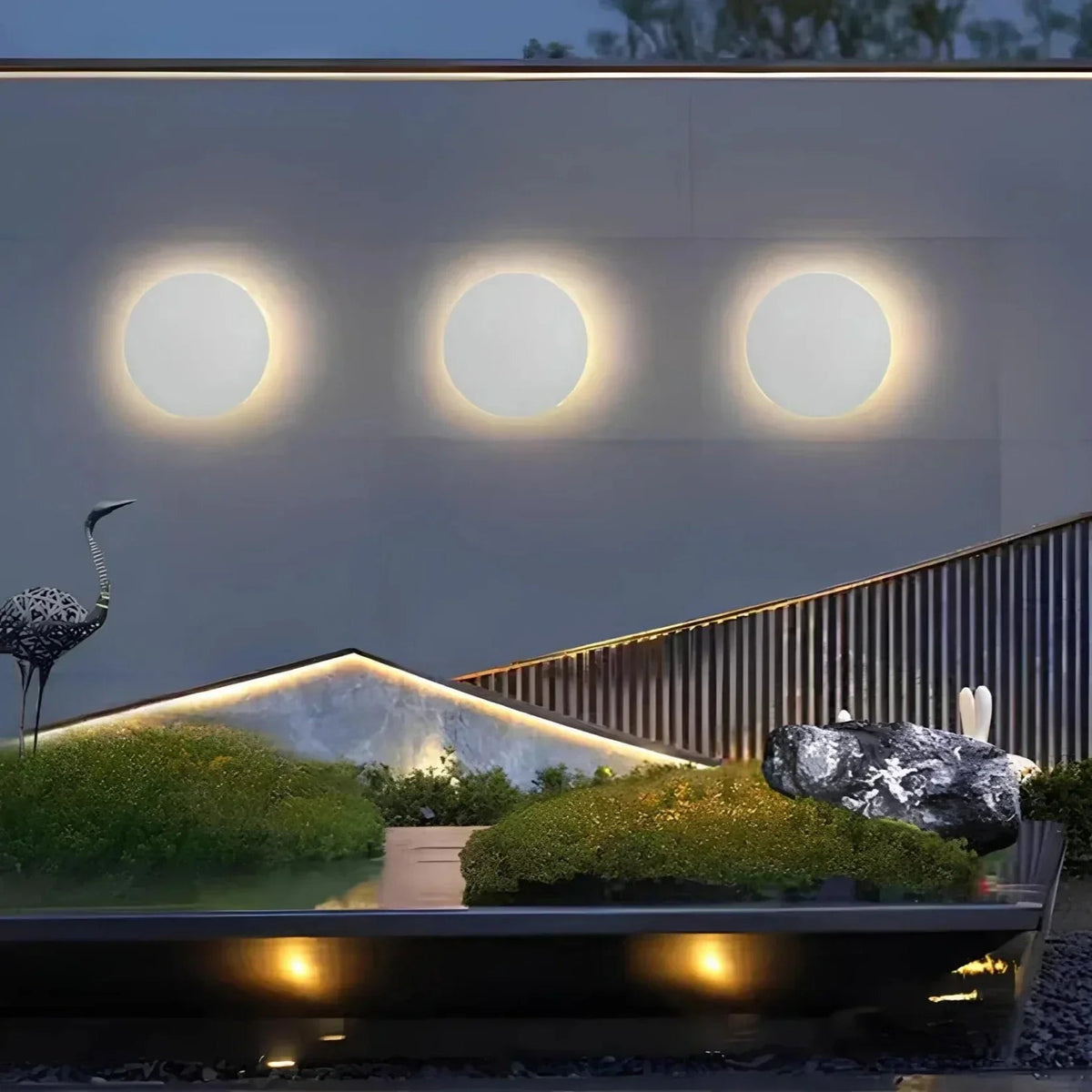 Drei gleich große weiße runde Wandleuchten in einer Reihe an einer Gartenmauer über einer modernen Landschaft mit Teich und Moos. 5