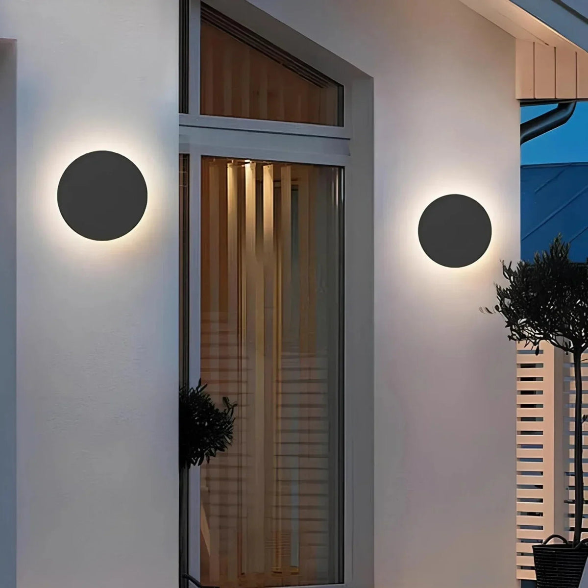 Zwei schwarze runde Wandleuchten an einer hellen Hausfassade neben einem Fenster, abends mit sanftem Lichtkranz eingeschaltet. 3