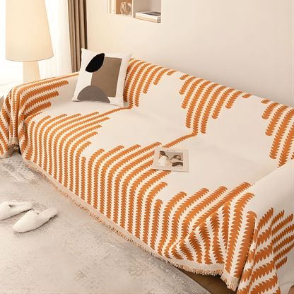 Schräge Ansicht eines Sofas mit orange-weiß gemustertem Überwurf und Fransenkante, dazu ein Kissen mit abstrakten Formen. 4