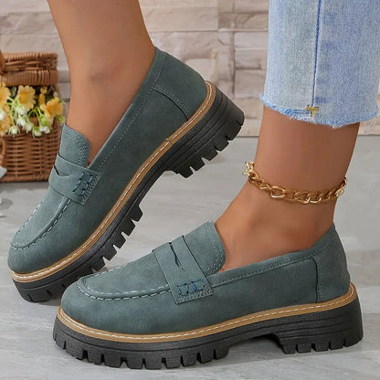 Graugrüne Loafer mit heller Naht und breiter Plateausohle an den Füßen einer Trägerin mit goldener Fußkette. 15
