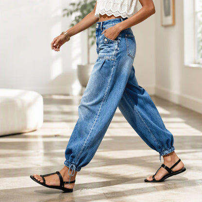Seitenansicht einer gehenden Frau in blauer Barrel-Jeans mit Handtasche, Saumkordeln und schwarzen Riemensandalen in einem hellen Raum. 7