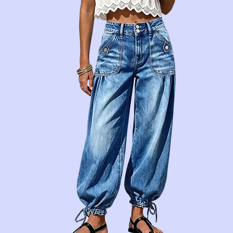 Nahaufnahme der Vorderseite: blaue Jeans mit hohem Doppelknopfbund, aufgesetzten Taschen mit Knopf und Raffung am Saum, vor fliederfarbenem Hintergrund. 10