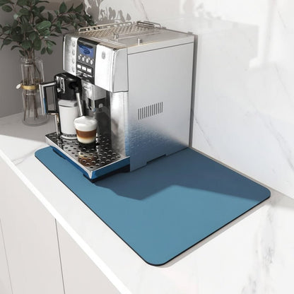 Blaue Unterlage mit abgerundeten Ecken unter einem Kaffeevollautomaten auf einer weißen Marmor-Arbeitsplatte, daneben eine Pflanze in einer Glasvase. 1
