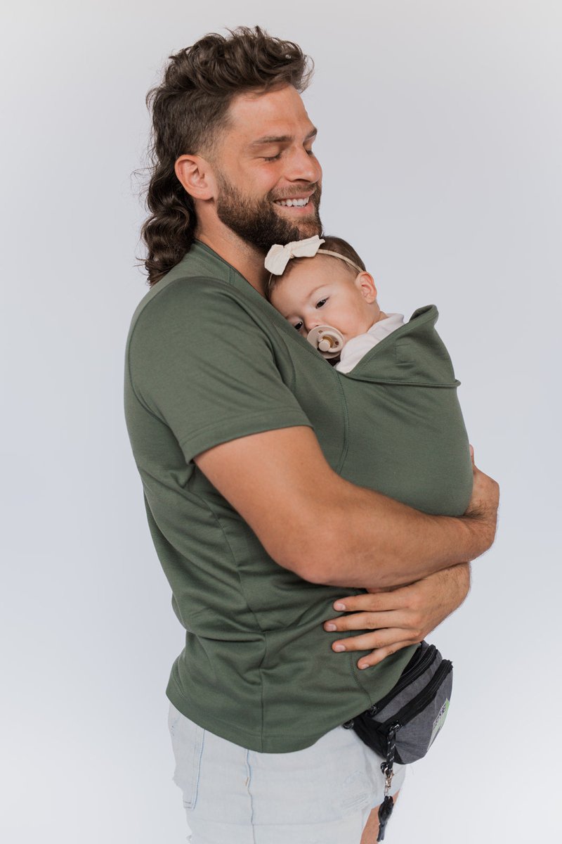Mann in grünem T-Shirt hält Baby in Tragetuch, lächelnd, weißer Hintergrund.